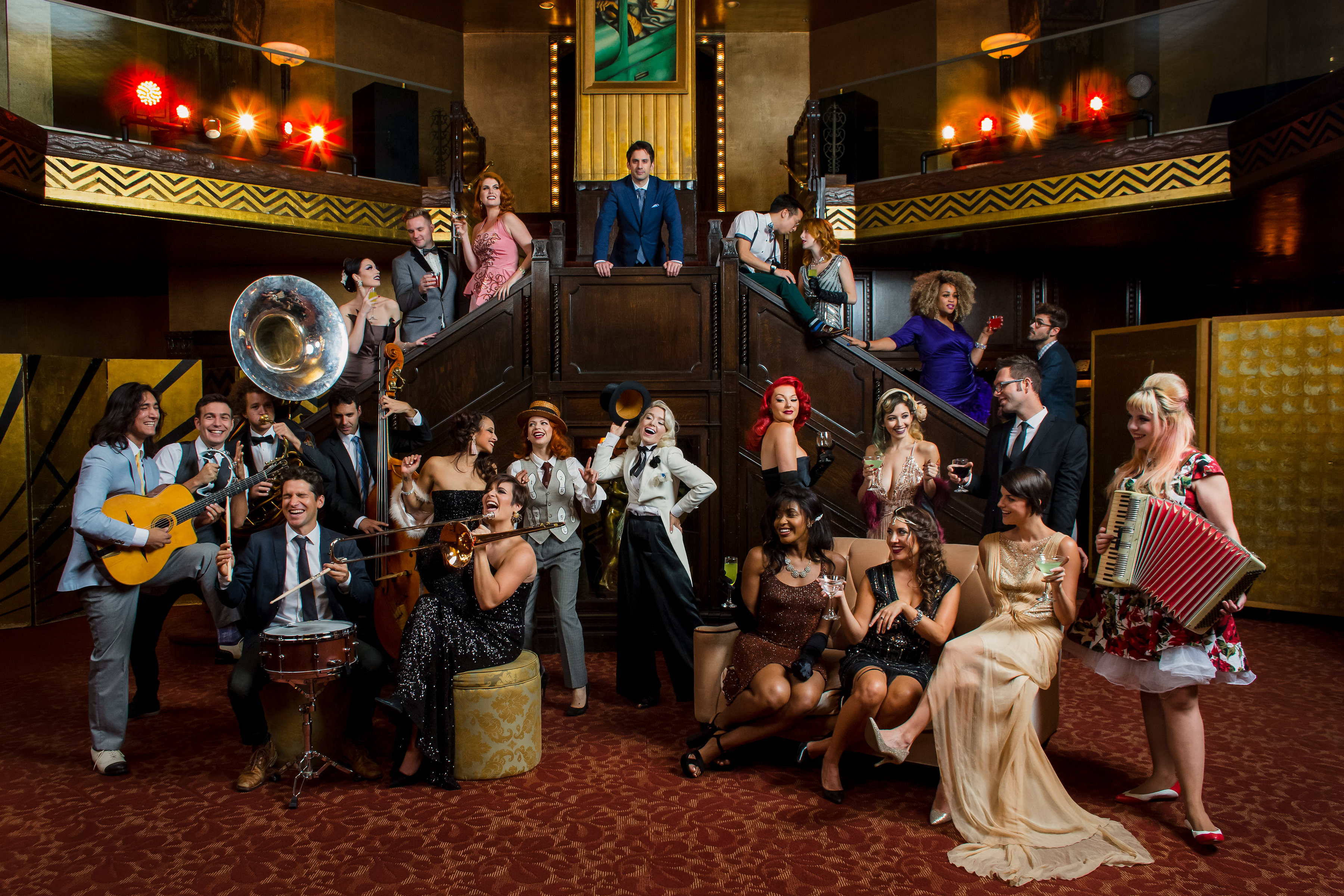 An image of the group Postmodern Jukebox. There are 25 people standing, laughing, and playing instruments in a midcentury modern speakeasy. The women are wearing colorful dresses and the men are wearing suits.