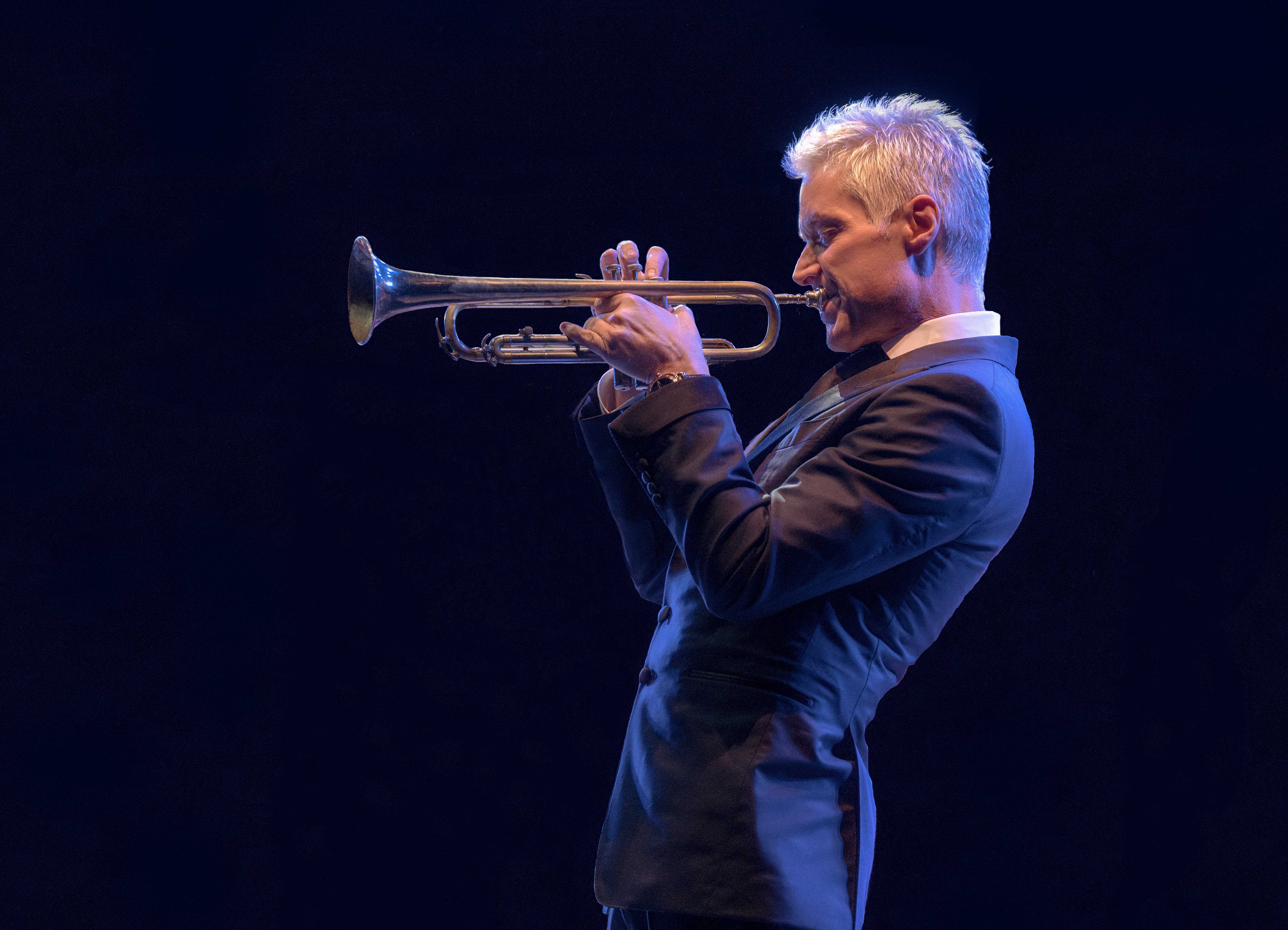 An image of Chris Botti. He is facing left, wearing a dark grey suit jacket and white button-up. His eyes are closed and he is playing the trumpet.